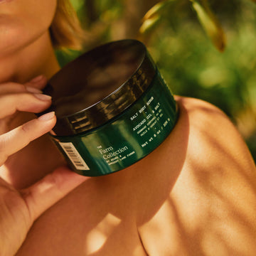 Hand holding a green jar labeled 'The Farm Collection' against a blurred natural background